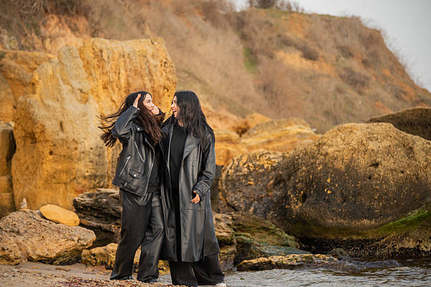 Tender moment between a mother and daughter embracing by the sea. Love, warmth, and family connection captured in a candid outdoor portrait. Windy coastal scene with genuine emotions and happiness.